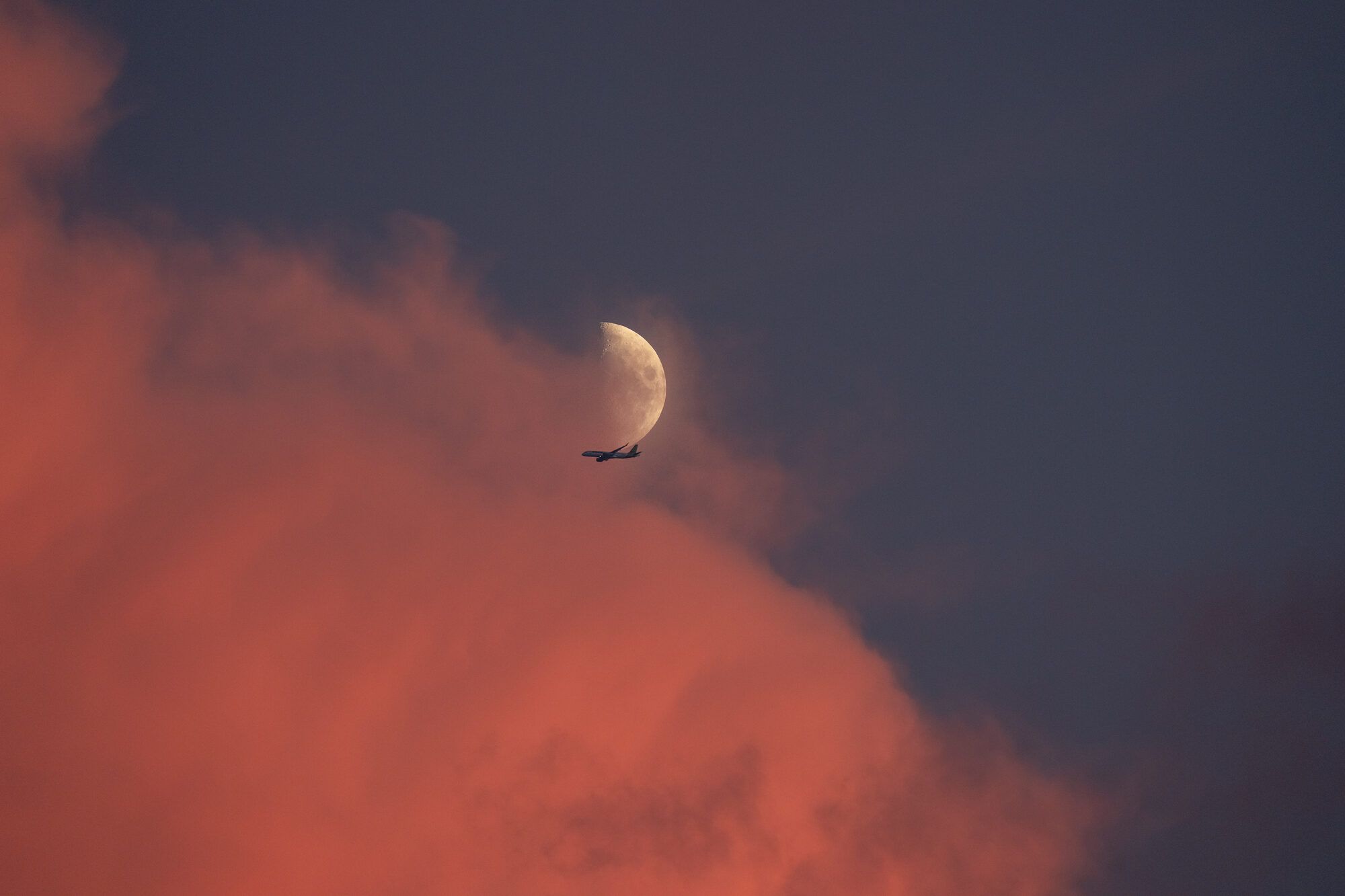 A waxing half moon above a silhouetted airliner, coral afterglow clouds below